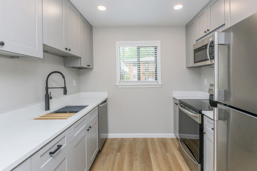 Modern kitchen featuring light gray cabinetry, a stainless steel refrigerator, a sleek oven, and a sink with a black faucet. A window with blinds allows natural light, and the floor is covered in light wood-style flooring.