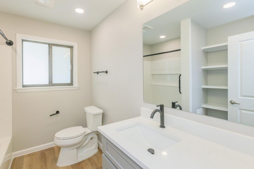 A modern bathroom featuring a white toilet, a sink with a contemporary faucet, a large mirror, and a window allowing natural light. The walls are painted light gray, and there is a simple shelving unit to the side.