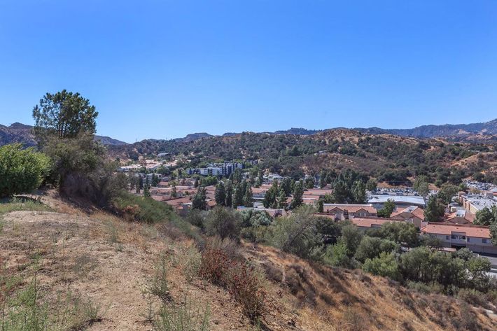 A panoramic view of a hilly landscape featuring residential buildings in the foreground, surrounded by greenery and mountains in the background under a clear blue sky.