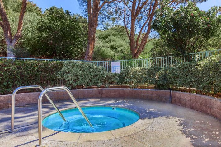 A small circular hot tub surrounded by greenery and plants, with a metal railing and steps leading into the water. The scene is set in a peaceful outdoor environment with trees in the background.