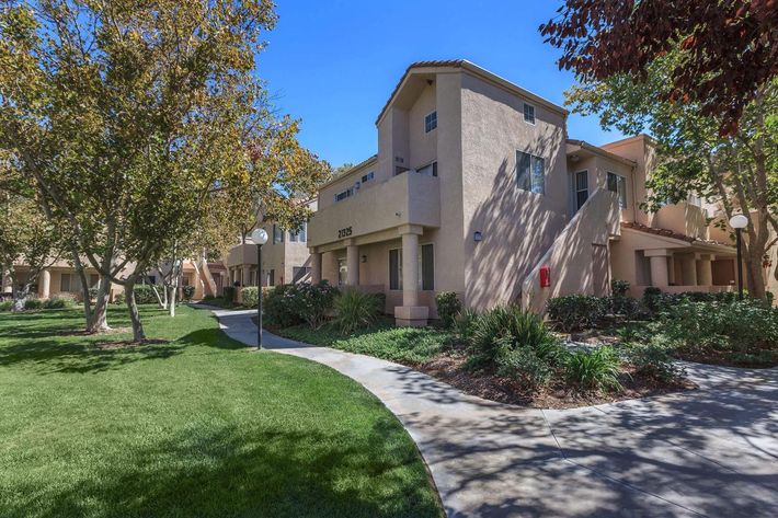 A pathway through a landscaped area with trees and shrubs leads to a residential building. The building features a light-colored exterior and is surrounded by green grass.