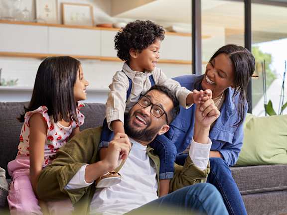 A family of four is sitting on a couch, with a child on the shoulders of the man.
