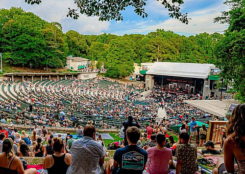 Vibrant outdoor amphitheater filled with an energetic crowd, surrounded by lush greenery under a clear sky.