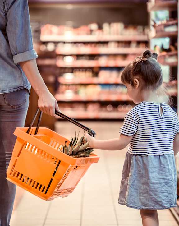 A woman and a little girl are shopping, the girl is holding a basket full of groceries.