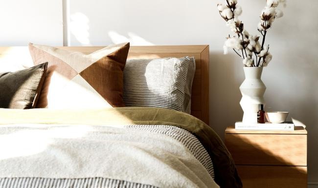 swath of sunlight shines across a well made bed and bedside table with a vase of cotton plants