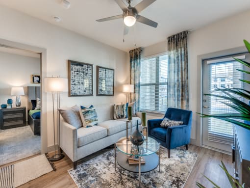 Luxury Apartments in Saginaw, TX, With In-Unit Washer And Dryer - Grand at Saginaw - Living Room With Sofa And Accent Chair, Glass Coffee Table, Ceiling Fan, And Balcony Door With Blinds.