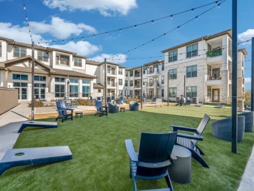 Pet-Friendly Luxury Apartments in Saginaw, TX - Grand at Saginaw - Courtyard Lawn With Adirondack Seating, String Lights Overhead, Cornhole Boards, And Surrounding Multi Story Apartment Buildings.