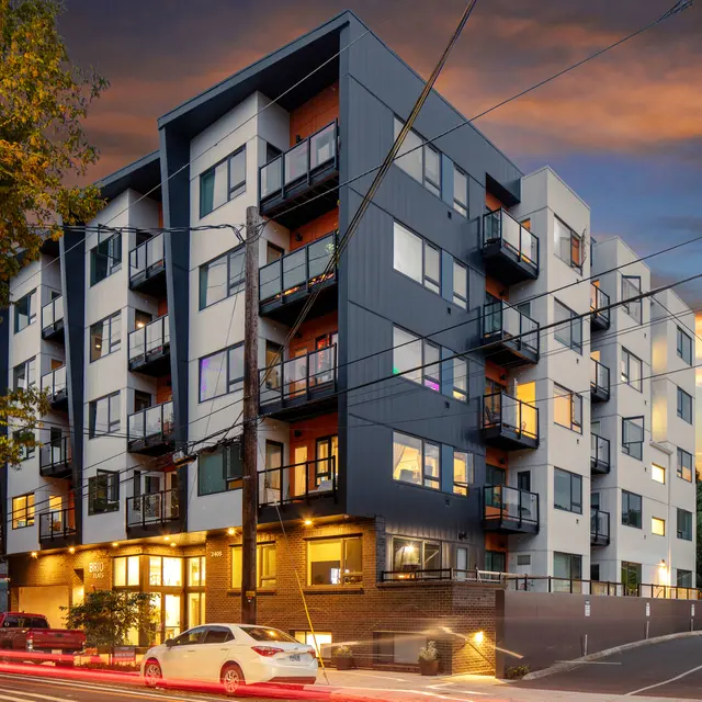 A modern multi-story apartment building with balconies, illuminated windows, and a sunset sky. A white car is parked near the building.