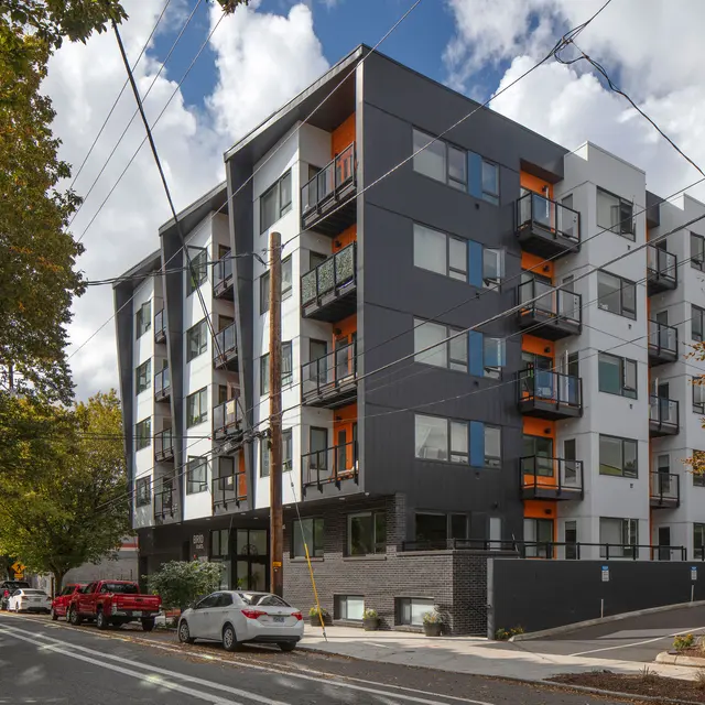 A contemporary multi-story apartment building with a mix of black and white exteriors and orange accents, situated along a tree-lined street with parked cars and a cloudy sky.