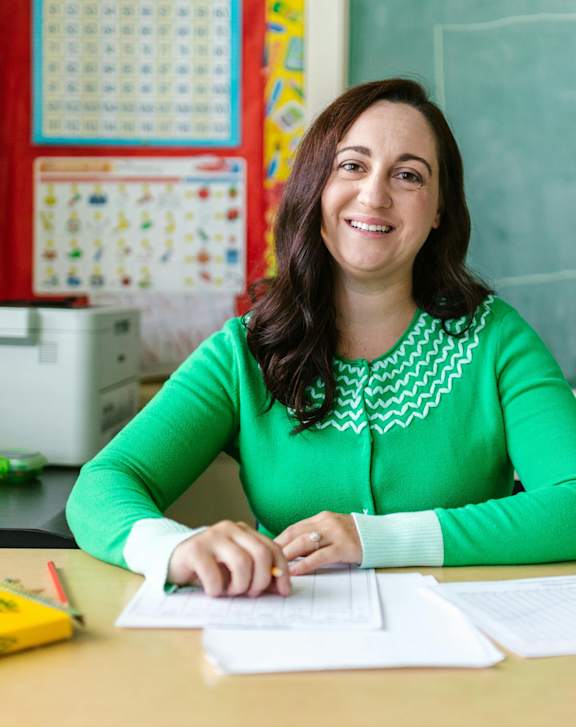 A woman in a green top is sitting at a desk with papers and a yellow box.