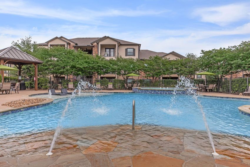 A vibrant swimming pool with two fountains creating splashes of water, surrounded by stone flooring. In the background, there are lush green trees and a multi-story building with a wooden gazebo, chairs, and umbrellas set up for relaxation. The sky is clear, adding to the inviting atmosphere.