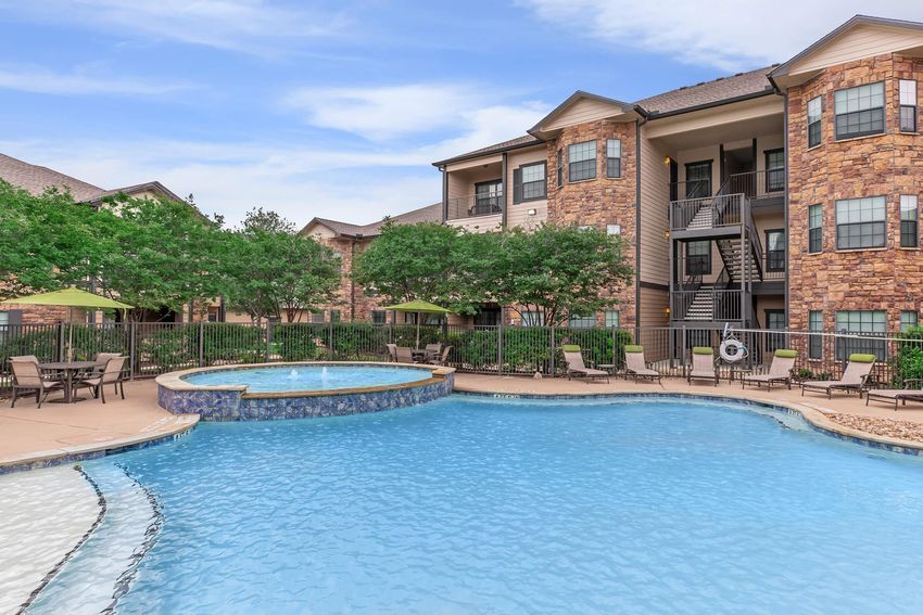A clear blue swimming pool surrounded by lounge chairs and tables, with green umbrellas. In the background, there are apartment buildings featuring stone and siding exteriors, and landscaped areas with small trees and shrubs. The scene is bright and inviting, under a clear blue sky.