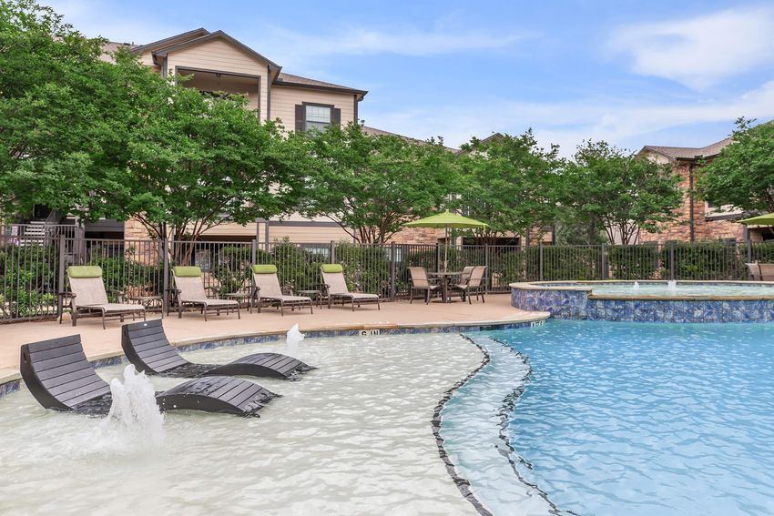 A serene outdoor swimming pool area featuring lounge chairs along the water's edge, surrounded by lush greenery. The pool has both a shallow section and a larger area, with water jets creating a relaxing atmosphere. In the background, there are apartment buildings with a warm, inviting design.