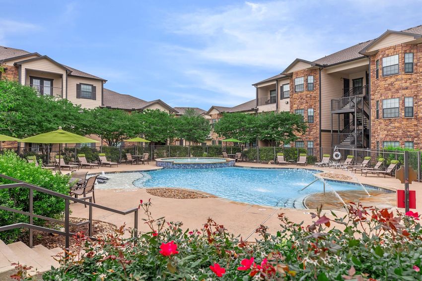 A landscaped pool area surrounded by apartment buildings. The pool features a curved design with a shallow area and is bordered by lounge chairs and umbrellas. Lush greenery and flowering plants enhance the setting, creating a relaxing outdoor atmosphere. Clear blue skies are visible above.