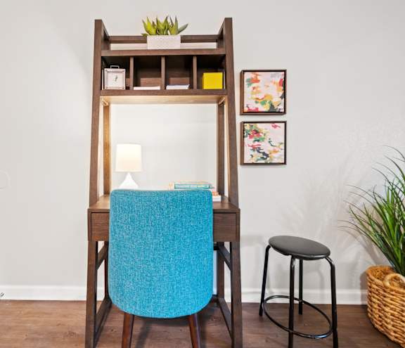 A blue chair sits in front of a wooden desk with a mirror and a lamp.