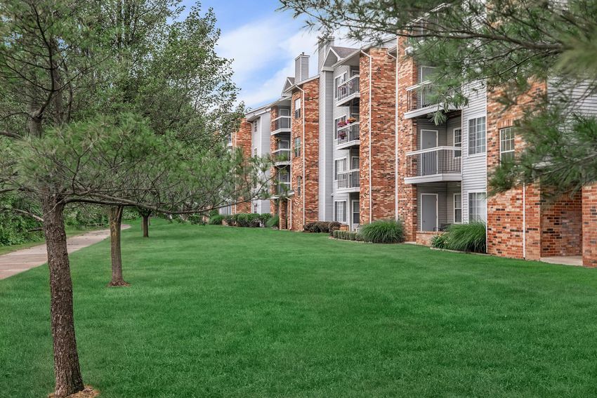 Apartment buildings with brick exteriors and balconies, surrounded by green grass and trees, under a blue sky.