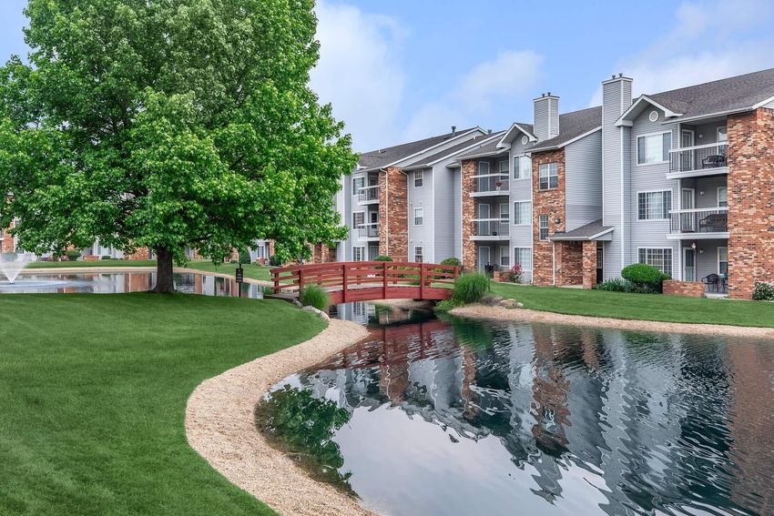 A serene apartment community with multiple buildings, featuring a green lawn, a small curved bridge over a tranquil pond, and lush trees surrounding the area. Reflections of the buildings and trees are visible in the calm water.