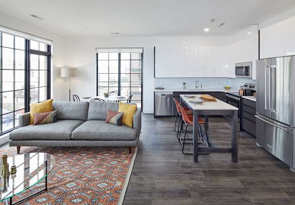 A modern apartment living space features a gray sofa with colorful pillows, a patterned rug, and a sleek kitchen with white and gray cabinets.