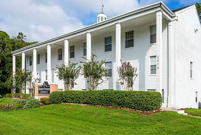 Elegant colonial-style apartment building with tall white pillars, lush landscaping, and a sunny curb appeal.