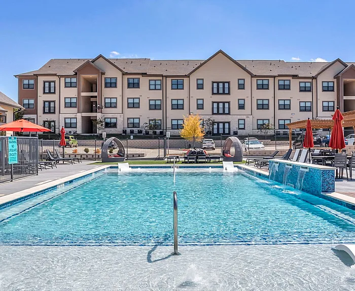 Luxurious apartment complex featuring a stunning outdoor pool, elegant lounge chairs, and vibrant red umbrellas under a clear blue sky.