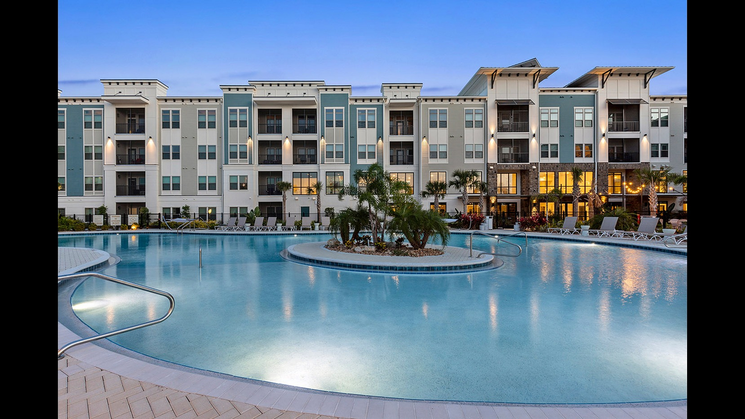 Luxurious apartment complex with modern architecture, featuring a stunning illuminated swimming pool surrounded by lush palm trees at dusk.