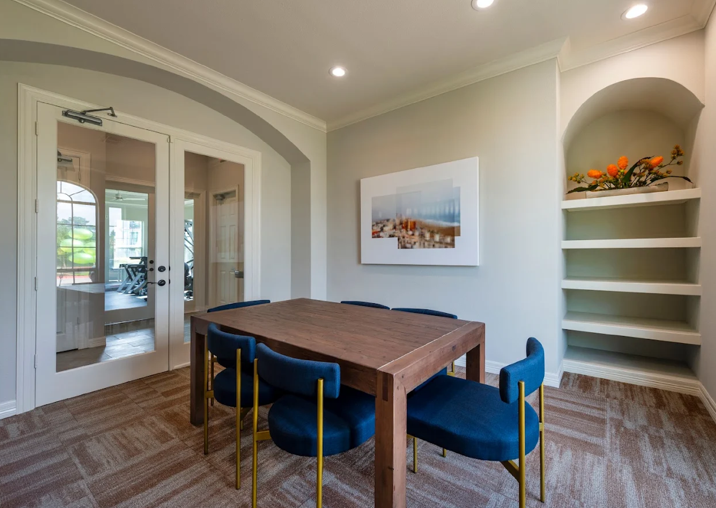 A modern dining room with a wooden table surrounded by six blue chairs with gold legs. There is a framed photo or painting on the wall, decorative orange flowers on a built-in shelf, and glass double doors leading to an exercise room with treadmill and weights visible inside.