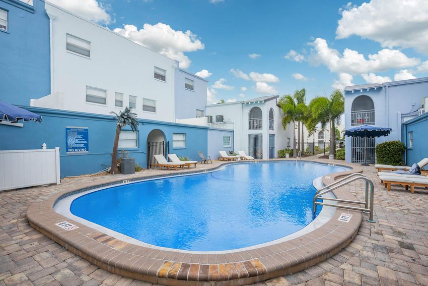 A serene swimming pool surrounded by a cobblestone patio, featuring lounge chairs and umbrellas. Bright blue skies with fluffy white clouds overhead, alongside palm trees and a light blue building that adds to the tranquil atmosphere. Ideal for relaxation and enjoyment.