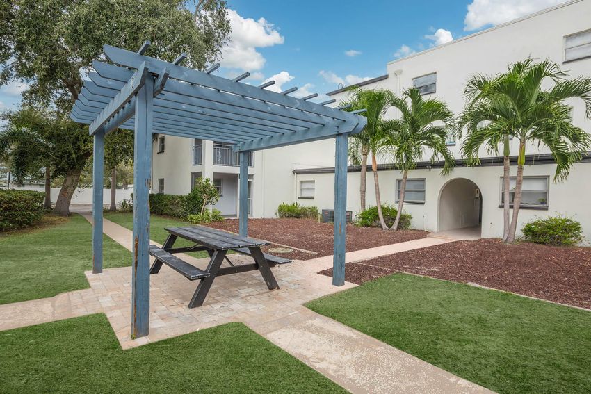 A wooden pergola with a picnic table underneath, surrounded by green grass and landscaping. In the background, there are white residential buildings and palm trees, with a clear blue sky above.