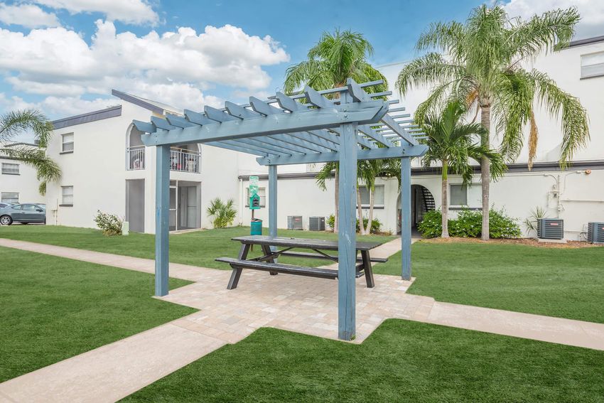 A wooden pergola with a picnic table underneath, surrounded by well-maintained green grass. In the background, there are residential buildings and palm trees under a partly cloudy blue sky. Pathways lead through the area, enhancing the landscaped appearance.