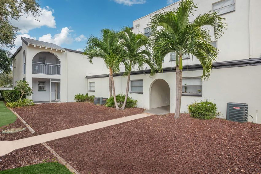 Exterior view of a building featuring two palm trees, a landscaped area with mulch, and air conditioning units. A walkway leads to the entrance, and the building appears to have multiple floors with balconies. The sky is partly cloudy, adding to the serene atmosphere.