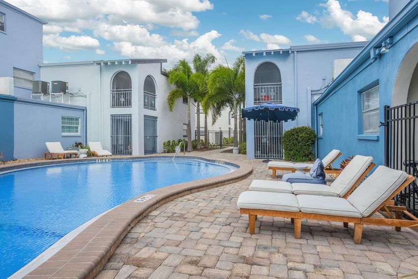 A serene swimming pool area featuring comfortable loungers, surrounded by palm trees and light-colored buildings. The clear blue water reflects the sky, while a canopy provides shade. The setting is inviting and ideal for relaxation.