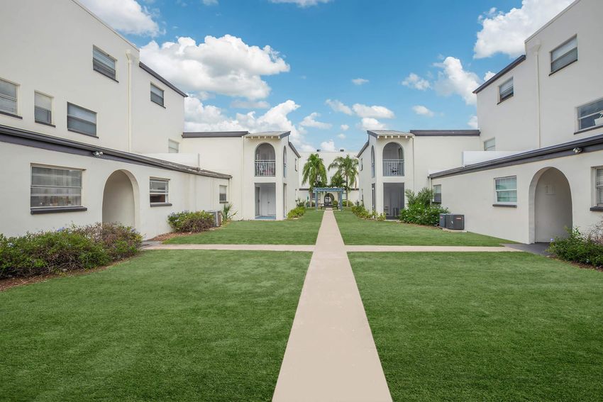 A well-maintained courtyard surrounded by white residential buildings. The green lawn features a clear pathway running through the center, leading to a small decorative archway and trees in the distance under a blue sky with fluffy clouds.