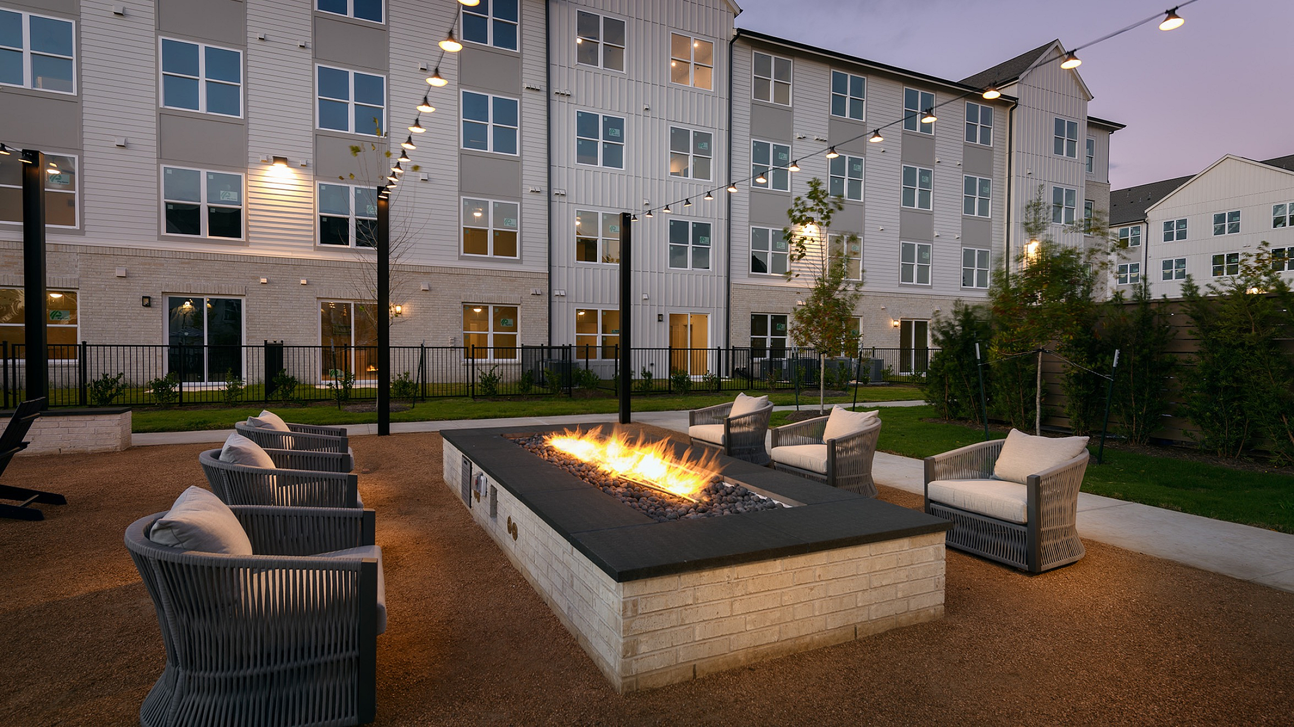 Charming outdoor lounge with a cozy fire pit surrounded by stylish seating, set against a modern apartment building at twilight.