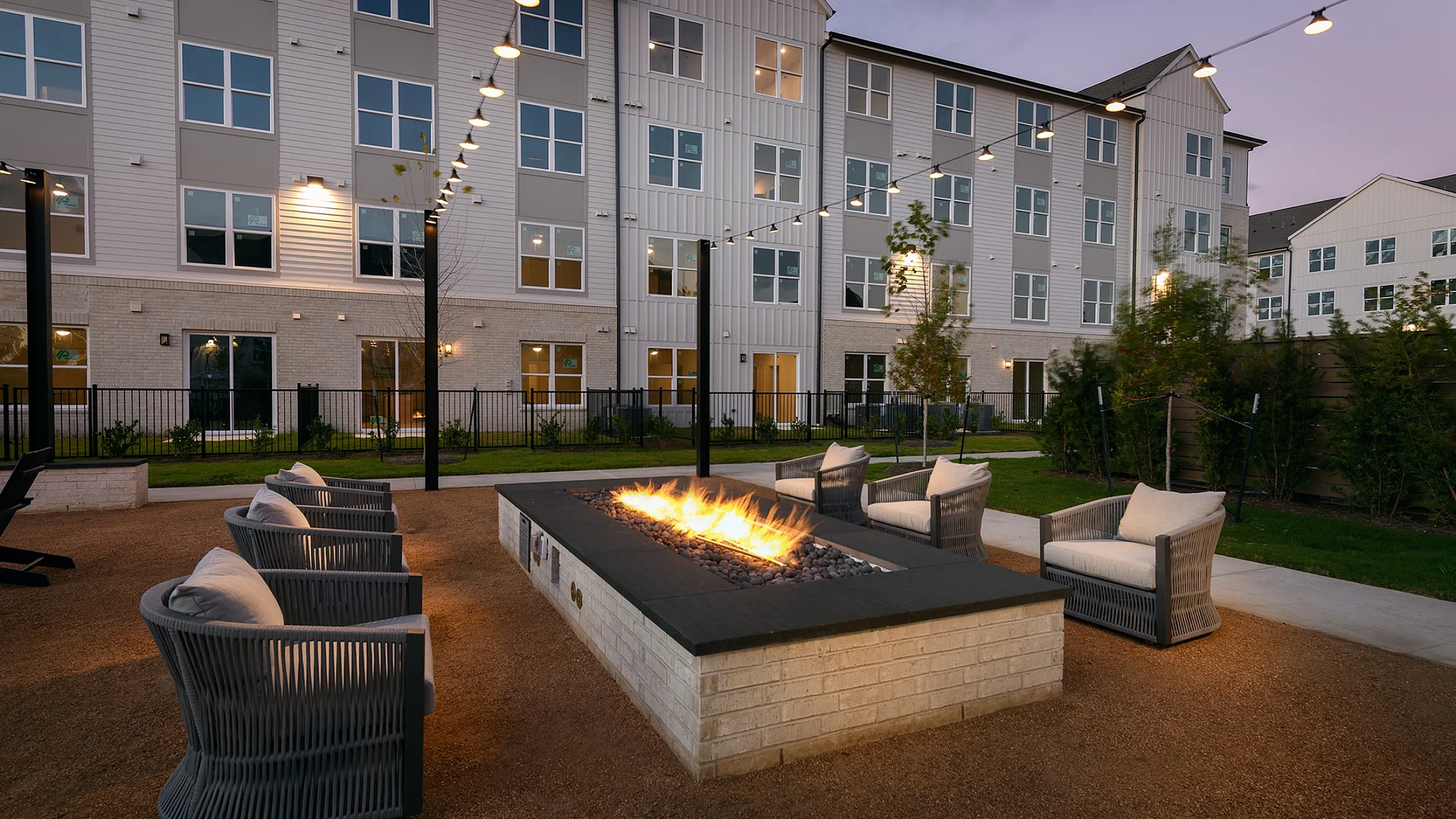 Charming outdoor lounge with a cozy fire pit surrounded by stylish seating, set against a modern apartment building at twilight.