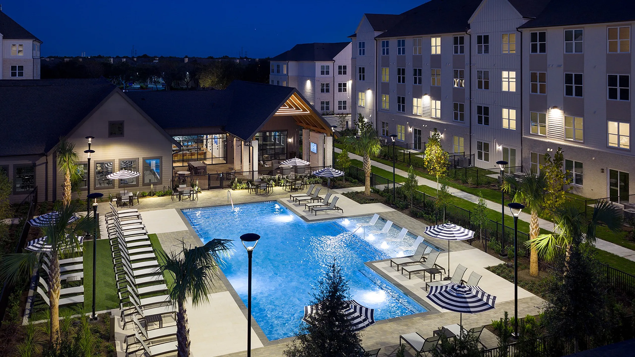 Elegant apartment complex with a beautifully lit swimming pool surrounded by lush landscaping and modern lounging areas, ideal for relaxing evenings.
