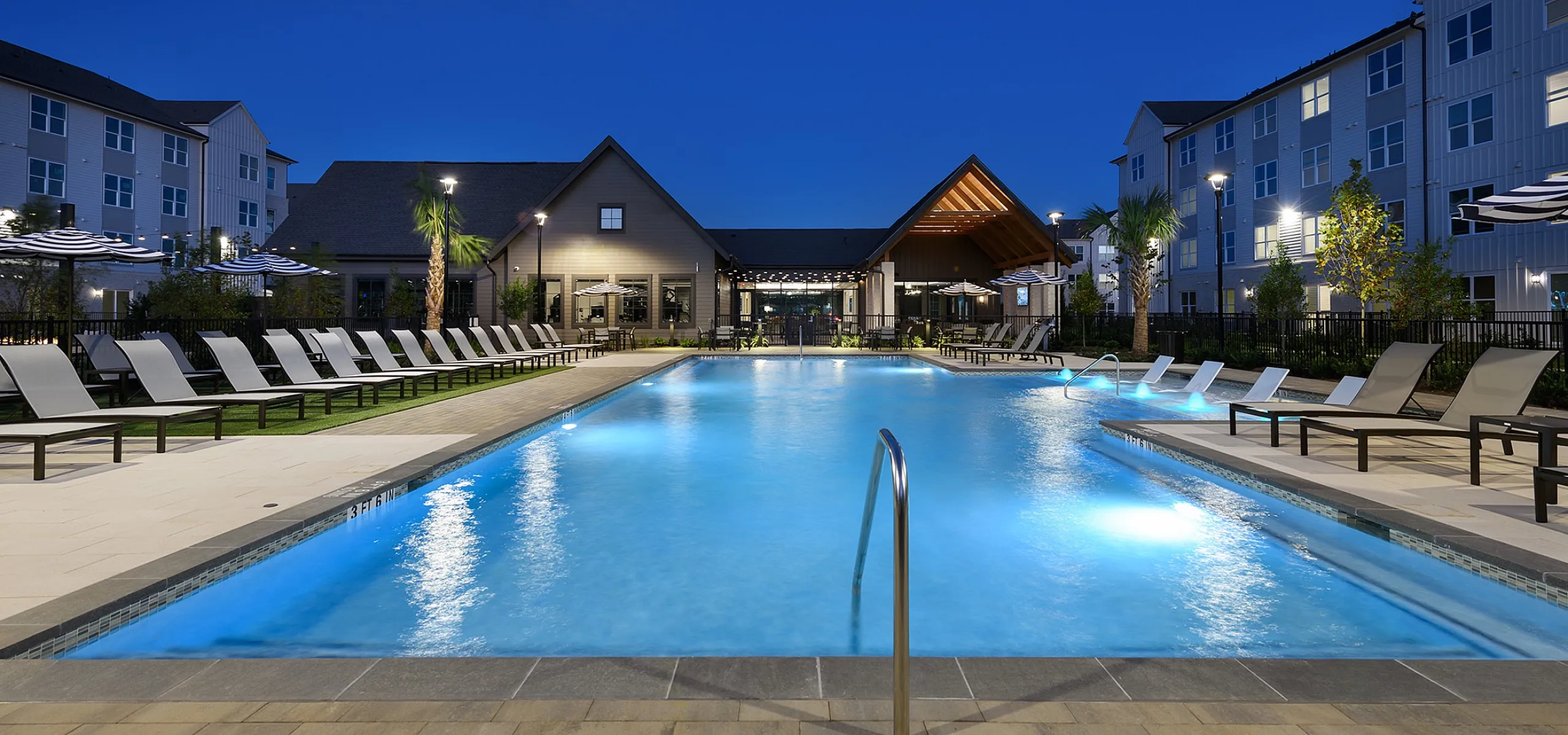 Luxurious apartment complex with inviting illuminated pool, surrounded by elegant lounge chairs and modern architecture under a stunning evening sky.
