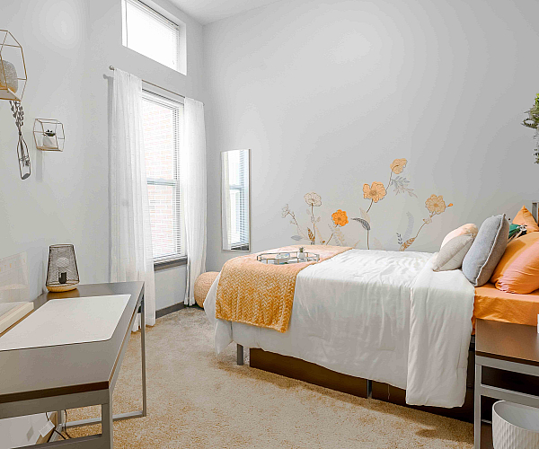 Charming bedroom with a cozy bed adorned with orange and white linens, decorative floral wall art, and modern geometric shelves. Bright natural light filters through the window, enhancing the serene and airy ambiance.