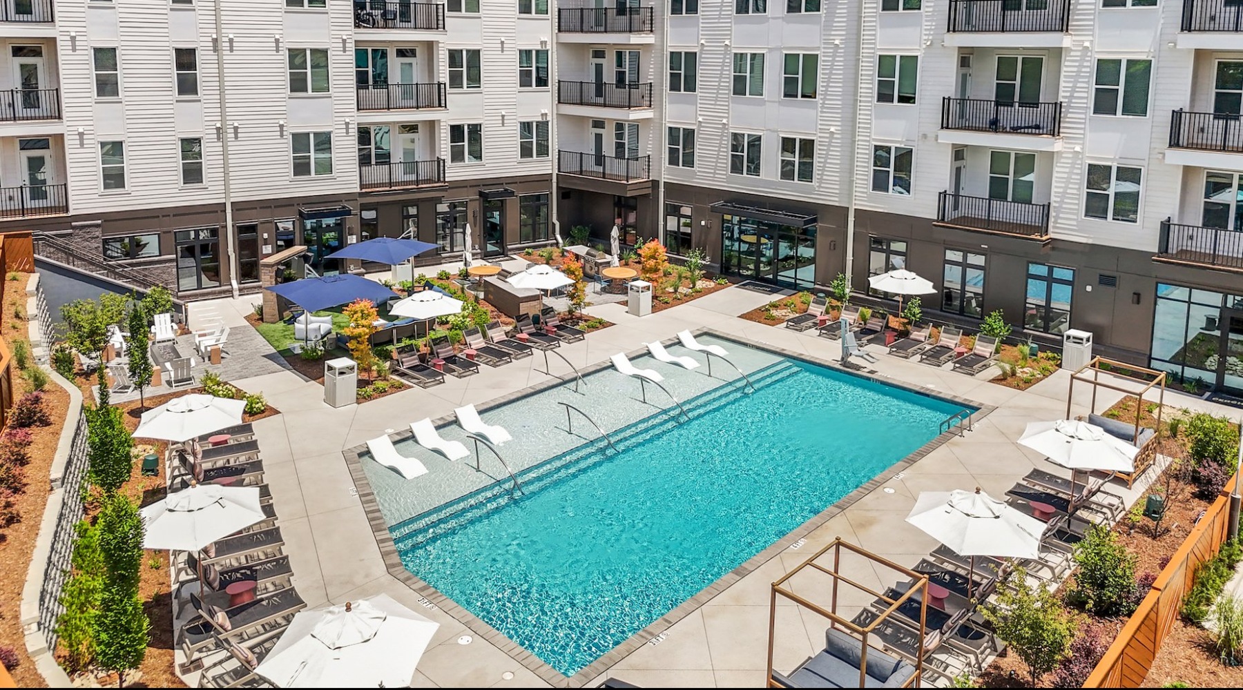 A large swimming pool surrounded by lounge chairs and umbrellas in front of a multi-story apartment building.