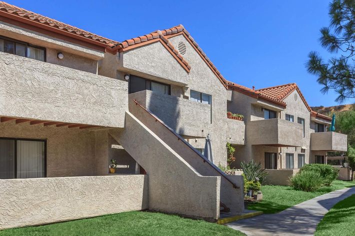 Row of beige apartment buildings with red tiled roofs, featuring balconies and a green lawn in front. Clear blue sky in the background.