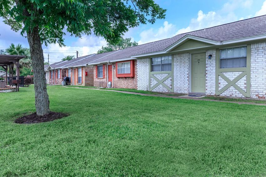 Row of duplex-style buildings with a grassy lawn in front, featuring trees and a cloudy blue sky overhead.