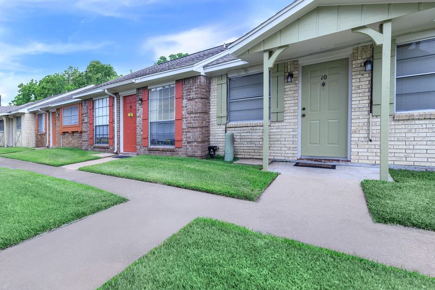 A row of residential buildings with red and beige brick exteriors, featuring green doors and large windows. The pathway in front is neatly paved, and the surrounding lawn is well-maintained with green grass.