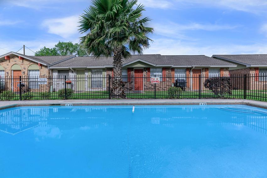 A clear blue swimming pool surrounded by a black fence, with a palm tree on one side and an apartment community in the background featuring red doors.