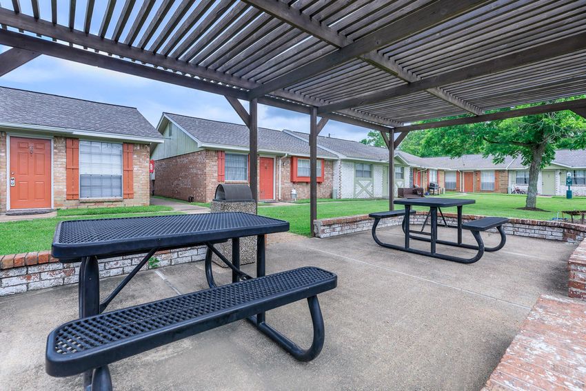 A shaded outdoor seating area with black picnic tables under a wooden pergola, surrounded by green grass and residential buildings in the background.