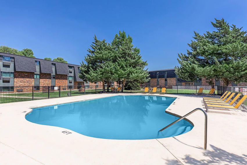 A clear blue swimming pool surrounded by a concrete deck, with yellow lounge chairs arranged nearby. In the background, a well-maintained apartment community with red brick and dark roofs is partially visible, along with several tall green trees under a bright blue sky.