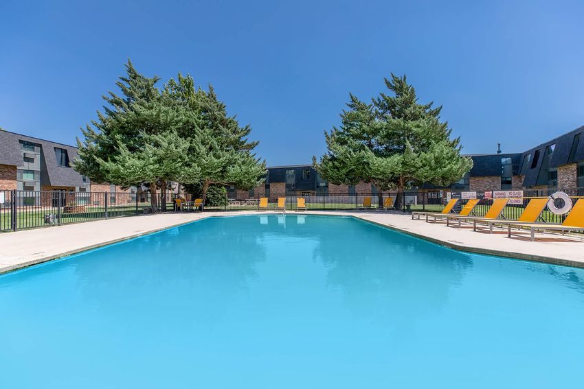 A clean, bright swimming pool surrounded by lounge chairs under a clear blue sky. Tall green trees flank the pool area, while modern buildings are visible in the background. The scene conveys a relaxing atmosphere, perfect for leisure and enjoyment.