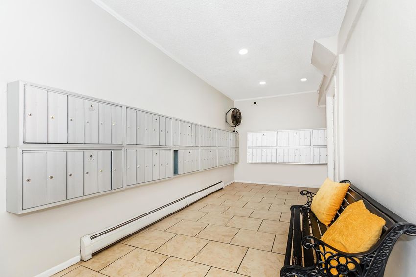 A hallway with a series of wall-mounted mailboxes along one side. There is a metal bench with two yellow cushions positioned in front of the mailboxes. The walls are painted light colors, and the floor is tiled. Ceiling lights provide illumination in the space.