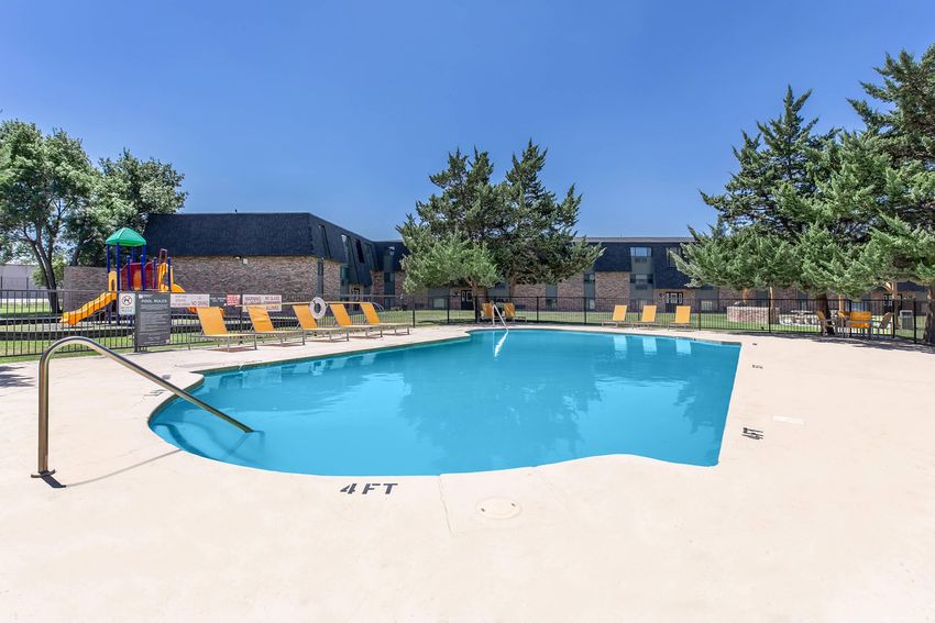 Swimming pool with clear blue water surrounded by a concrete deck. Yellow lounge chairs line one side of the pool, and a safety railing leads into the water. In the background, there are trees and apartment buildings. The area is sunny with a clear blue sky. Play structure visible on one side.