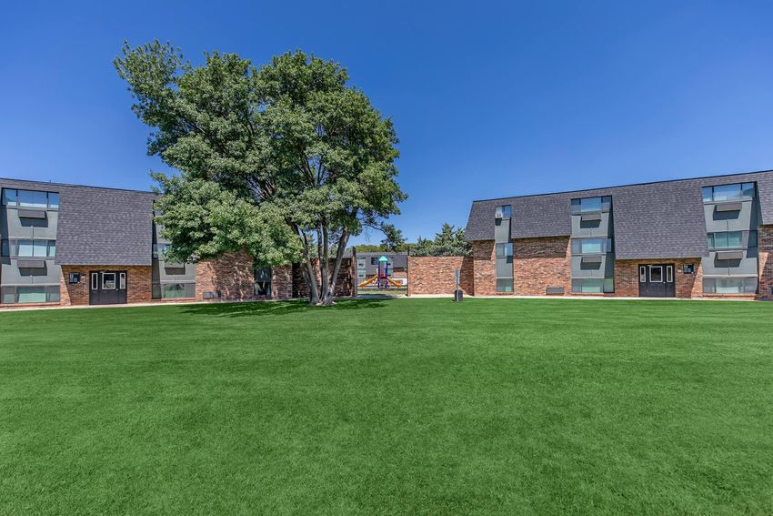 A well-maintained green lawn surrounded by two-story brick apartment buildings under a clear blue sky. A large tree is positioned centrally, providing shade, while the buildings display modern architecture with multiple entryways. The scene portrays a pleasant residential atmosphere.