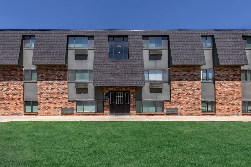 A multi-story apartment building with a brick and gray exterior. The building features large windows, a central entrance with double doors, and air conditioning units below the windows. A well-maintained grassy area is in front of the building under a clear blue sky.
