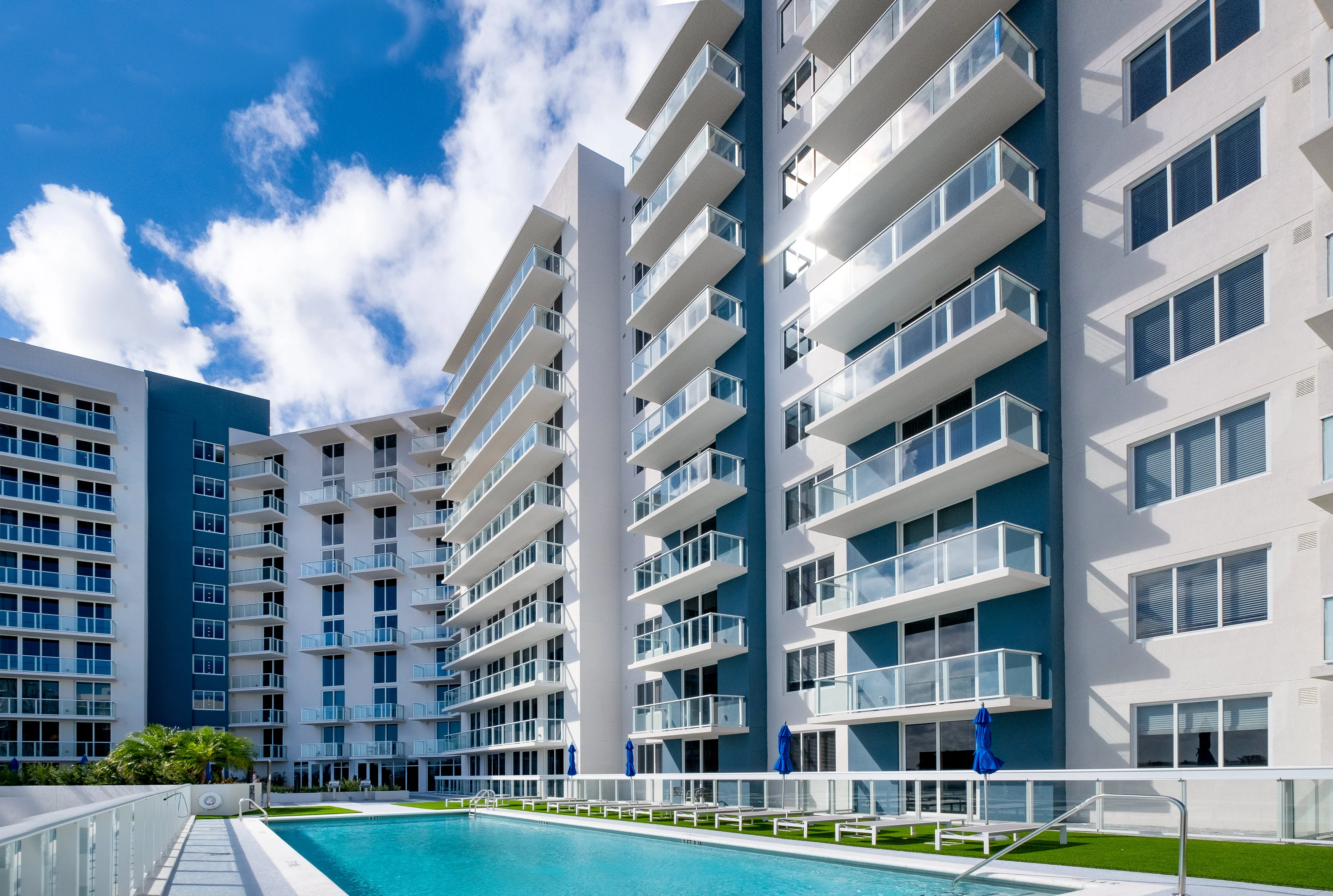 A modern apartment complex featuring multiple buildings with balconies and a swimming pool in the foreground under a blue sky.